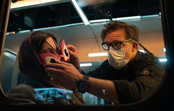 A man in glasses and a white mask adjusts large pink cartoon eyes on a person wearing a red hood.