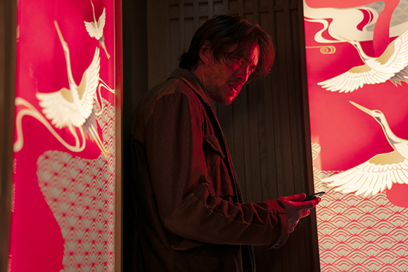 A man in a brown jacket looks down at a smartphone between two red panels featuring white crane illustrations.