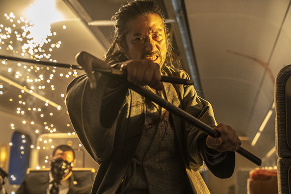 A man with long hair and a beard holds a cane and a long staff in a dark, industrial-looking room.