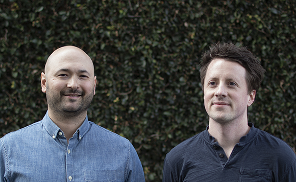 A bald man in a blue shirt and a man with dark hair in a dark shirt stand before a leafy hedge.