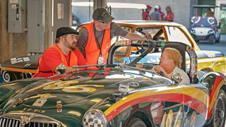 Pit crew members in orange vests talk to a driver seated in a green vintage racing car.