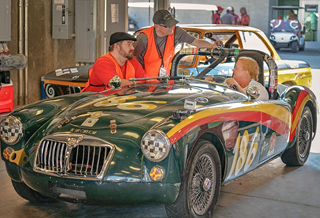 Mechanics in orange vests lean over a green vintage racing car with number 186 in a garage.