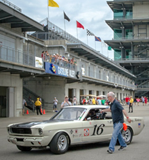 A white vintage Mustang race car with the number 16 drives past a concrete pit lane and grandstand.
