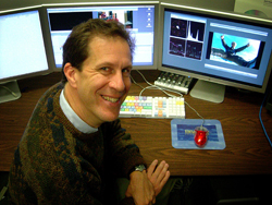 A man in a patterned sweater smiles while sitting at a desk with three computer monitors.