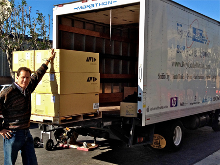 A man stands beside a large white moving truck with several yellow Avid boxes stacked on the open liftgate.