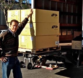 A man in a patterned sweater stands beside a tall stack of yellow Avid boxes on a pallet.