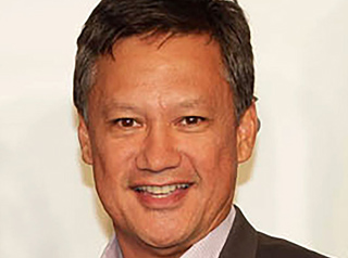 A smiling man with short dark hair wearing a collared shirt and a dark blazer.