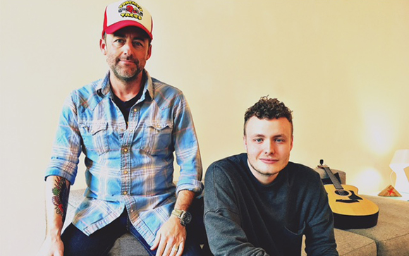 A man in a red cap and plaid shirt sits next to a man in a dark sweater on a couch.