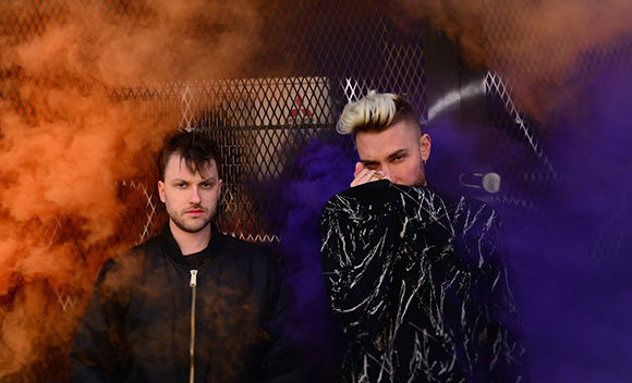 Two men stand behind clouds of orange and purple smoke against a wire mesh background.