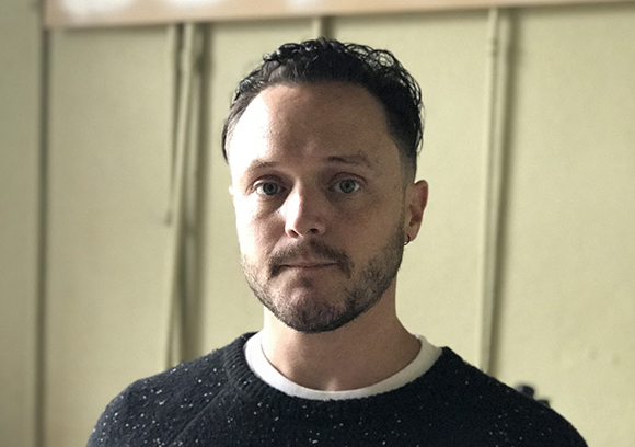 A man with short dark hair and a beard wearing a black speckled sweater.