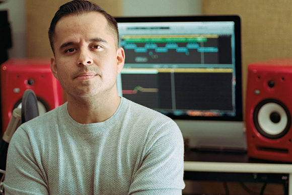 A man in a light blue sweater sits in a studio with red speakers and a computer monitor in the background.