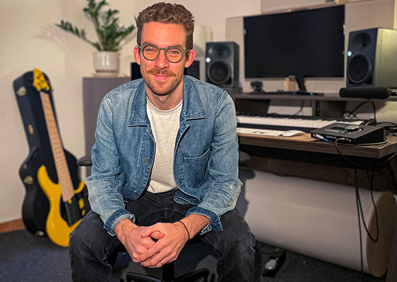 A man with glasses and a denim jacket sits in a music studio with a guitar case and computer equipment.