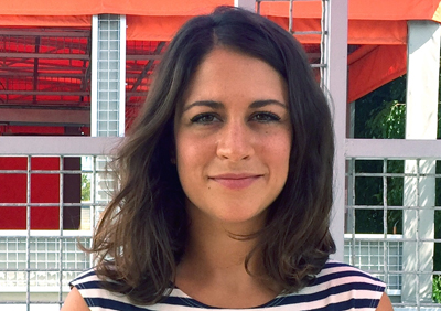 A woman with wavy brown hair and a striped shirt smiles in front of a red and white structure.