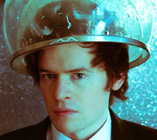 A man with dark hair wearing a clear, dome-shaped plastic hair-drying hood against a blue glittery background.