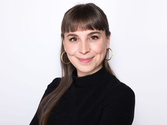 A smiling woman with brown hair, bangs, and gold hoop earrings wearing a black turtleneck.