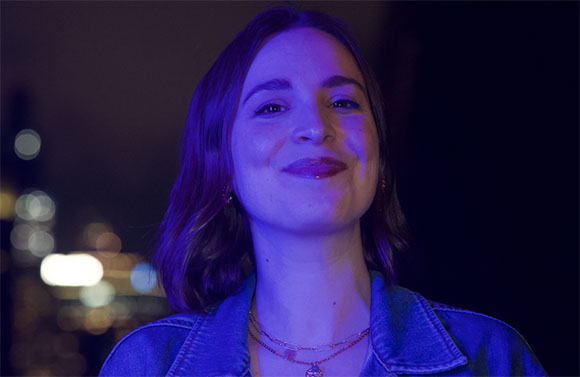 A smiling woman with shoulder-length hair illuminated by purple light against a dark, blurred background.