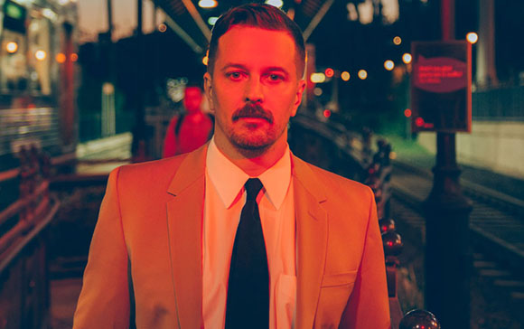 A man with a mustache wearing a tan suit and black tie stands on a train platform at night.