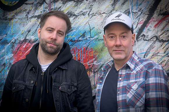 Two men pose in front of a vibrant, multi-colored graffiti-covered wall.