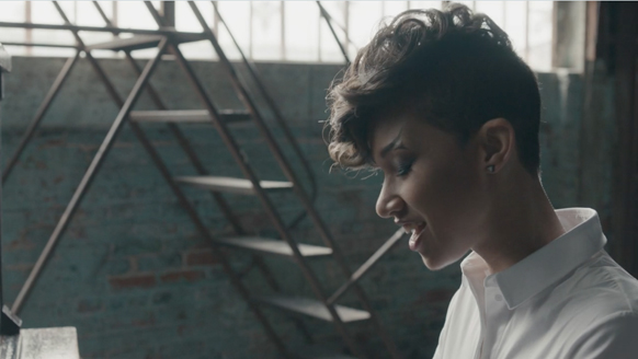 Profile of a person with short dark hair smiling in a dimly lit industrial space with a metal staircase.