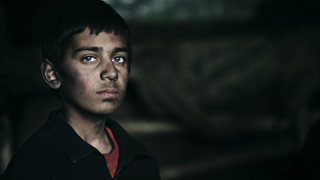 A young boy with dark hair and a somber expression looks toward the camera against a dark, blurred background.