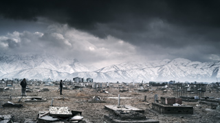 A vast, barren cemetery with scattered graves under a dark, stormy sky and snowy mountains.