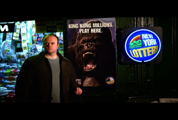 A man in a brown jacket stands next to a King Kong Millions poster and a New York Lottery sign.