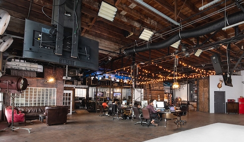 Industrial studio with high ceilings, exposed pipes, string lights, and workstations with computers.