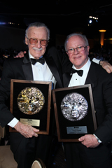 Two men in black tuxedos and bowties smiling while holding large circular commemorative plaques.