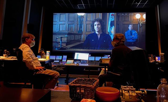 A person in a mask sits in a dark room watching a large screen displaying a man in a blue shirt.