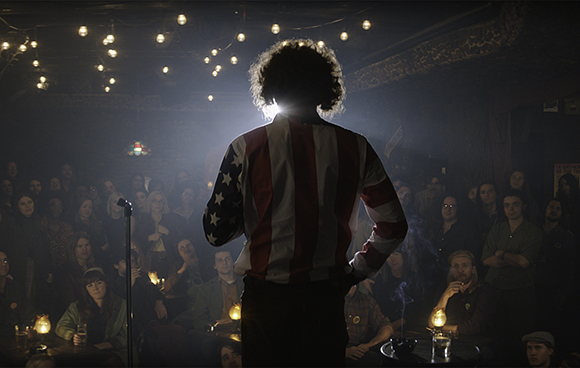 A person wearing a shirt with the American flag pattern stands with their back to the camera facing a crowd.
