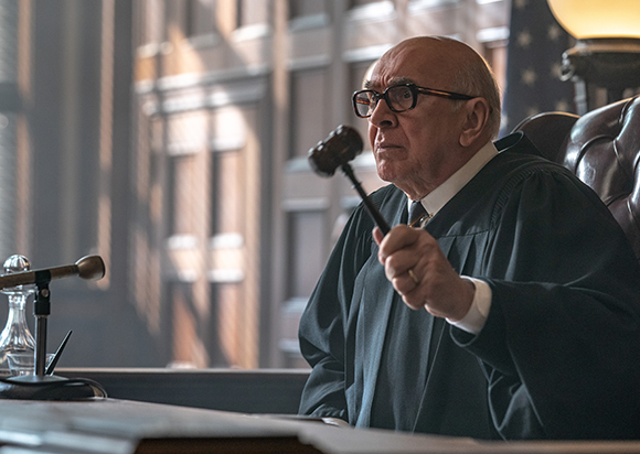 An older man wearing glasses and black judicial robes holds a wooden gavel in a courtroom.