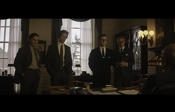 Four men in dark suits stand in a room with large windows and a desk in the foreground.