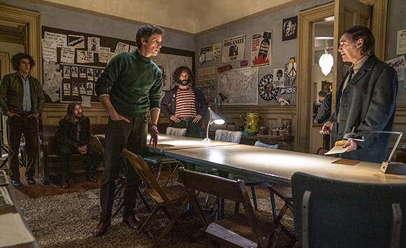 A man in a green sweater leans over a long table toward a man in a dark suit in a room filled with posters.