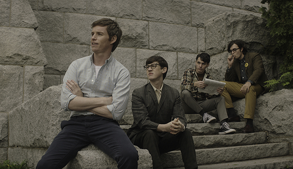 Four men in vintage-style clothing sit on large stone steps in front of a stone wall.