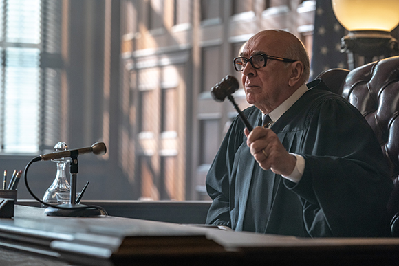 An elderly judge in black robes holds a wooden gavel in a courtroom setting.