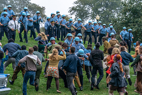 A crowd of people in blue uniforms and colorful vintage clothing running across a grassy slope.