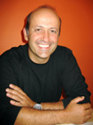 A smiling man with short dark hair and a black long-sleeved shirt against an orange background.