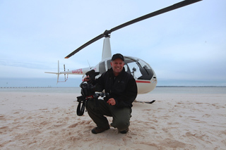 A man in a black jacket and cap kneels on a sandy beach holding a camera in front of a white helicopter.