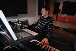 A man wearing glasses and a striped sweater plays a MIDI keyboard in a music studio.