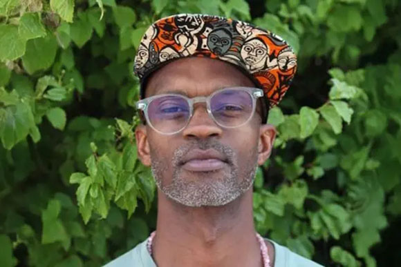 A man with a salt-and-pepper beard wearing a patterned cap and clear glasses against a background of green leaves.