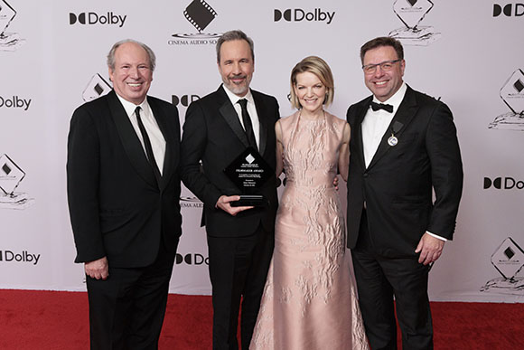 Four people in formal attire pose on a red carpet in front of a white backdrop with Dolby logos.