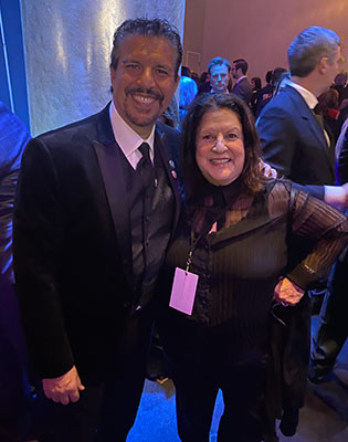 A smiling man in a black suit and a woman in a dark patterned blouse posing together at an indoor event.