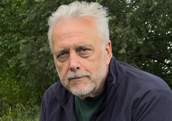 A close-up portrait of an older man with white hair and a grey beard against a background of green trees.
