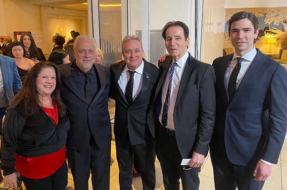 Five people in formal attire pose together for a group photo at an indoor event.
