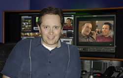 A man smiling in a studio with multiple video monitors displaying footage behind him.