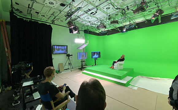 A film studio featuring a large green screen, studio lights, cameras, and crew members.
