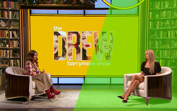 Two women sit in white armchairs facing each other on a brightly colored talk show set.