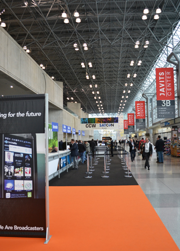 A wide view of a convention hall with a high metal truss ceiling, orange carpeting, and Javits Center banners.