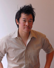 A man with dark, messy hair wearing a tan button-down shirt looking toward the camera.