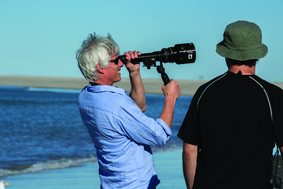 A man in a blue shirt looks through a large spotting scope on a tripod near the ocean.